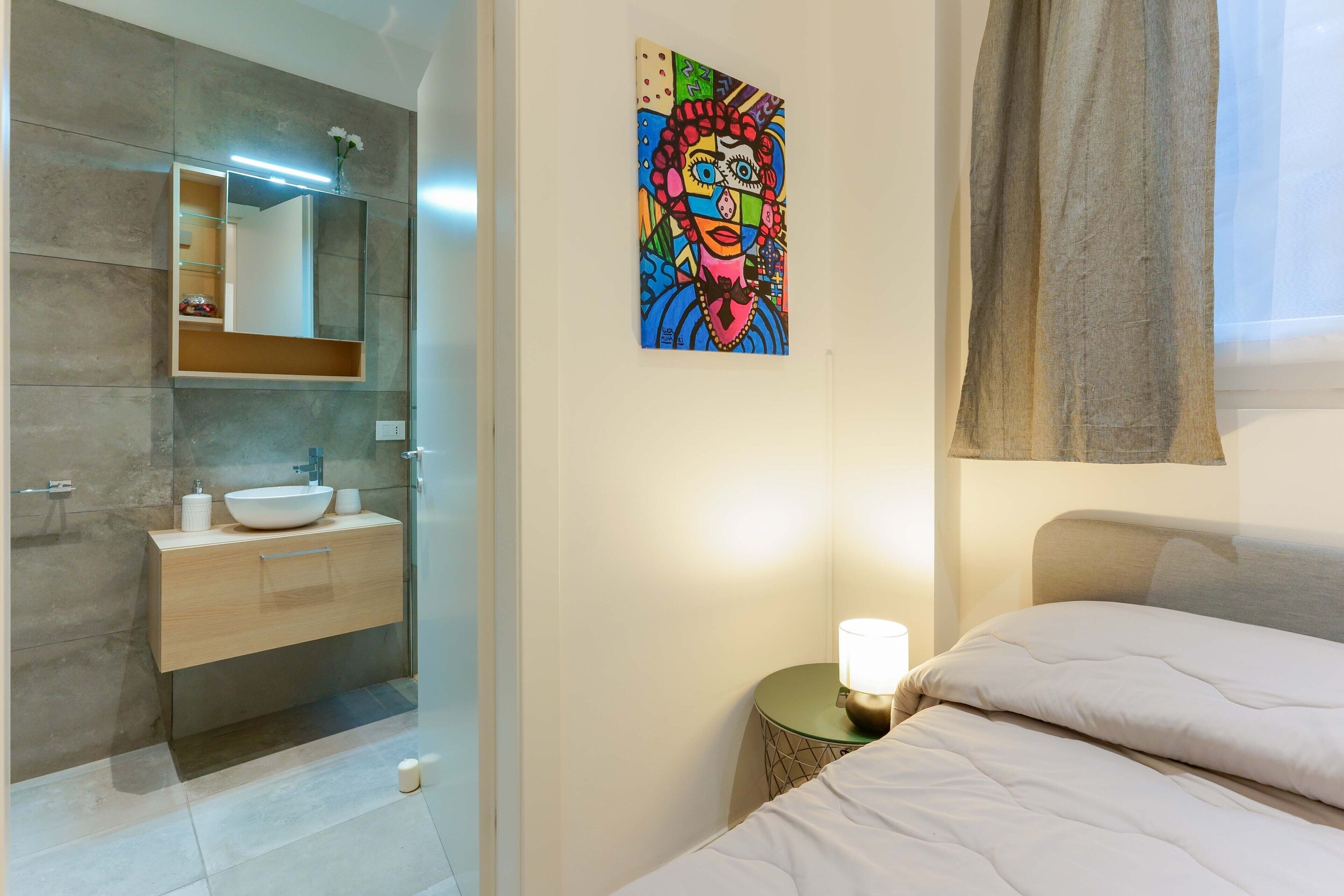 A modern bathroom with a glass-enclosed shower, a white sink, and a bedside table with a lamp.