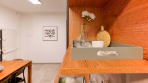 Un interior de madera con una estantería que sostiene un jarrón con flores, un recipiente de vidrio y una taza. Un cuadro enmarcado está en la pared.