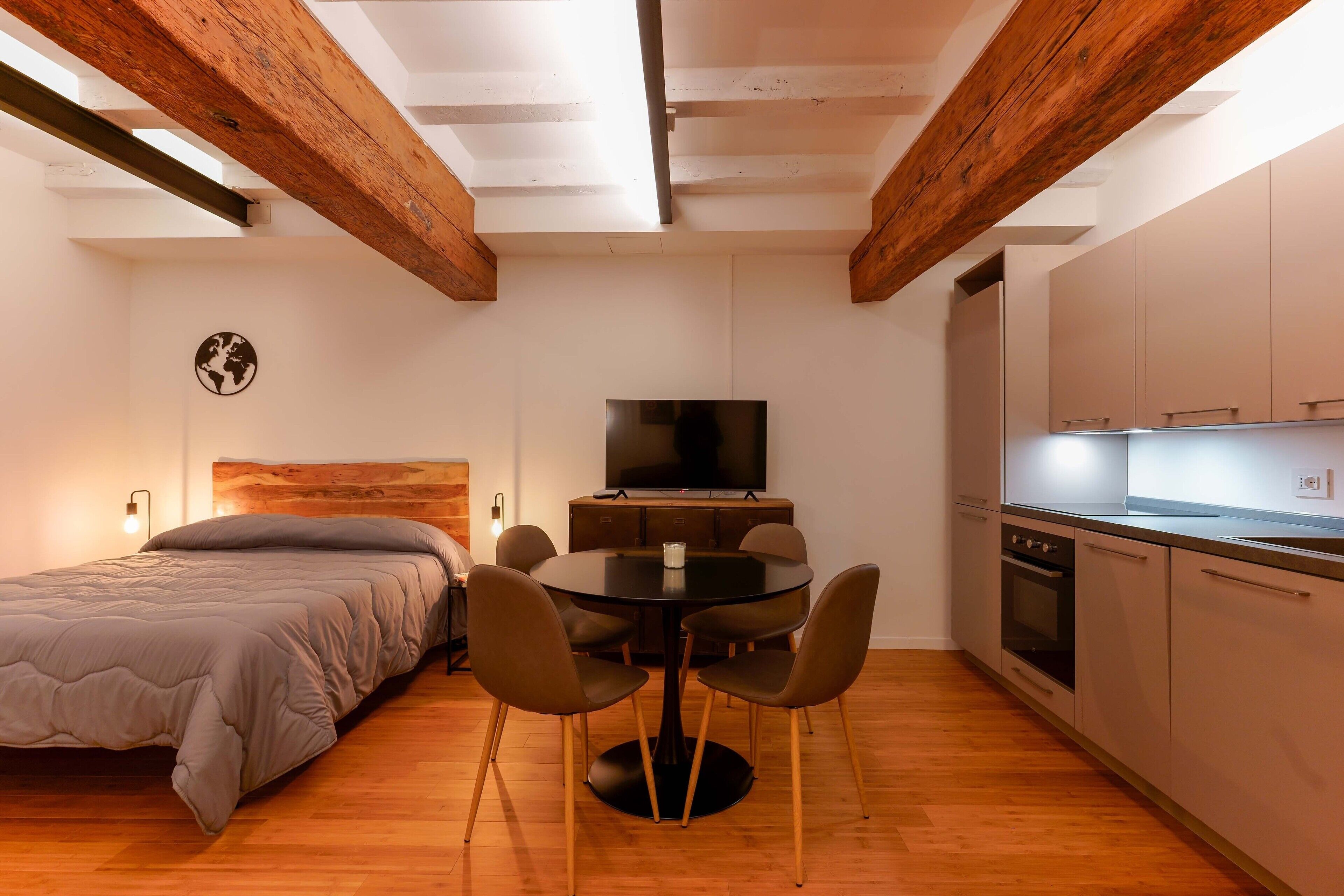 A compact room with a bed, a dining table with chairs, a TV, and a kitchenette.