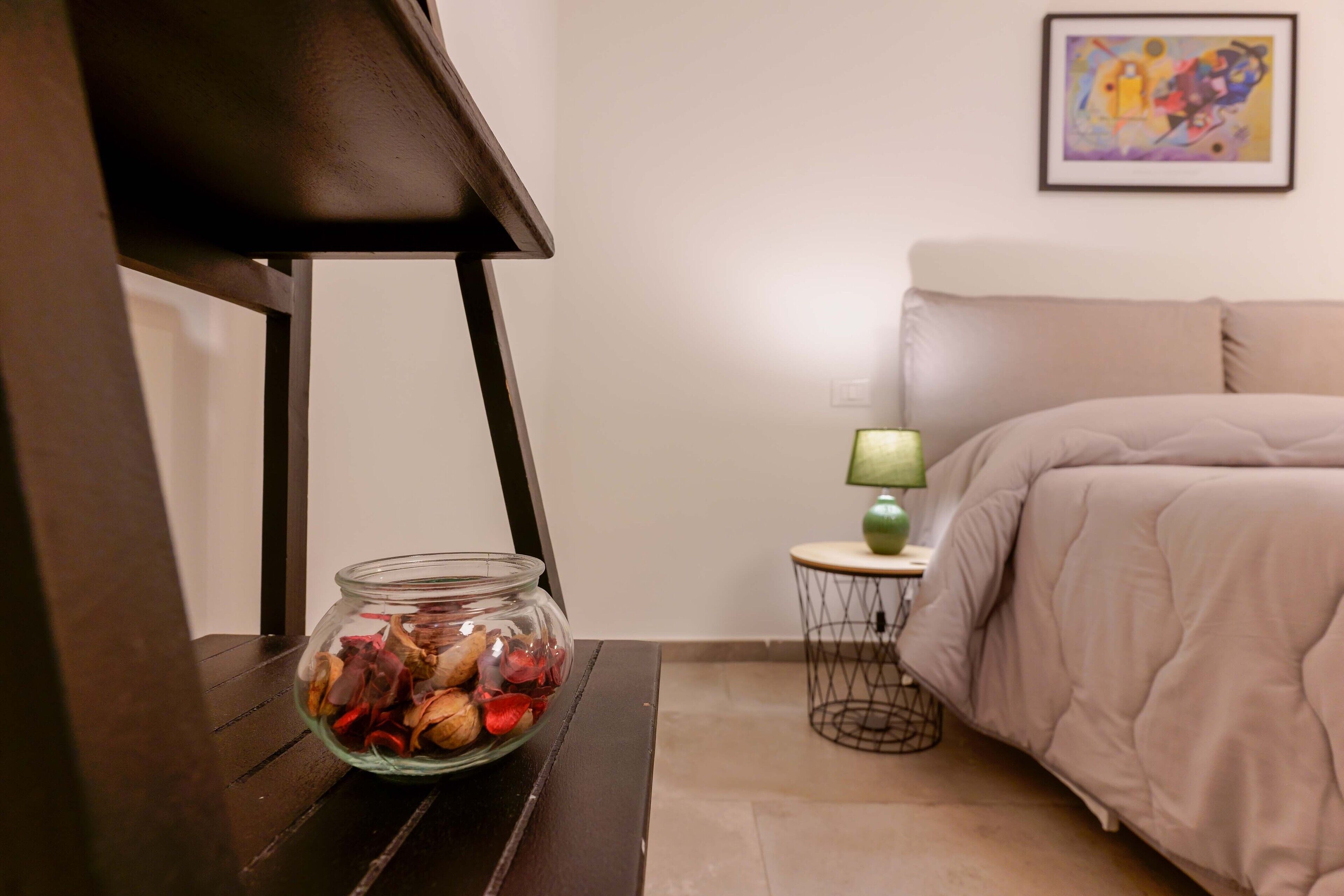 Ein Zimmer mit einem Bett, einer Lampe, einem kleinen Tisch und einem Glasbehälter mit Gegenständen darin.