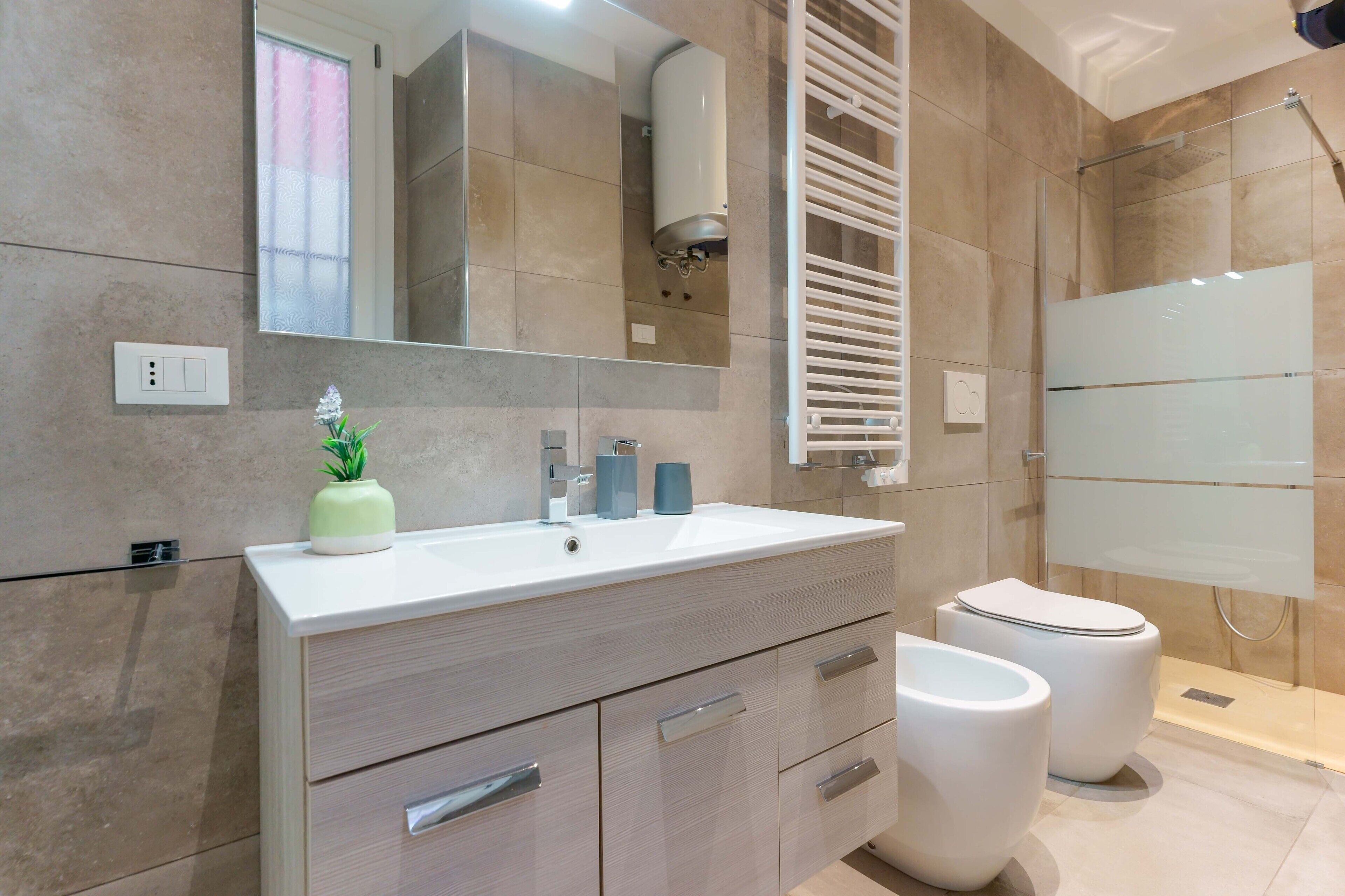 A modern bathroom with a beige tiled wall, a white vanity with a mirror, two toilets, and a glass shower enclosure.