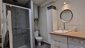 A modern bathroom with a glass shower enclosure, a toilet, a sink with a mirror, and a washing machine. - Le T2 A du Meysse (Meysse)
