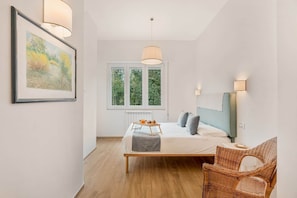 A bedroom with a bed, bedside table, lamp, and a wicker chair. - COLOMBO APARTMENTS (Lido di Camaiore)