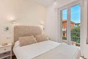 A bedroom with a large bed, two bedside tables, a window with a view of buildings, and a balcony. - COLOMBO APARTMENTS (Lido di Camaiore)