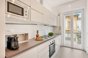 A modern kitchen with a gas stove, microwave, and oven, a coffee maker, and a view of a building through a glass door. - COLOMBO APARTMENTS (Lido di Camaiore)