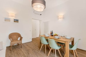 A dining area with a wooden table set for four, surrounded by chairs, and a wicker chair in the corner. - COLOMBO APARTMENTS (Lido di Camaiore)