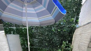 Een terras met een stenen plantenbak, twee witte stoelen, een gestreepte parasol en een plant in pot.