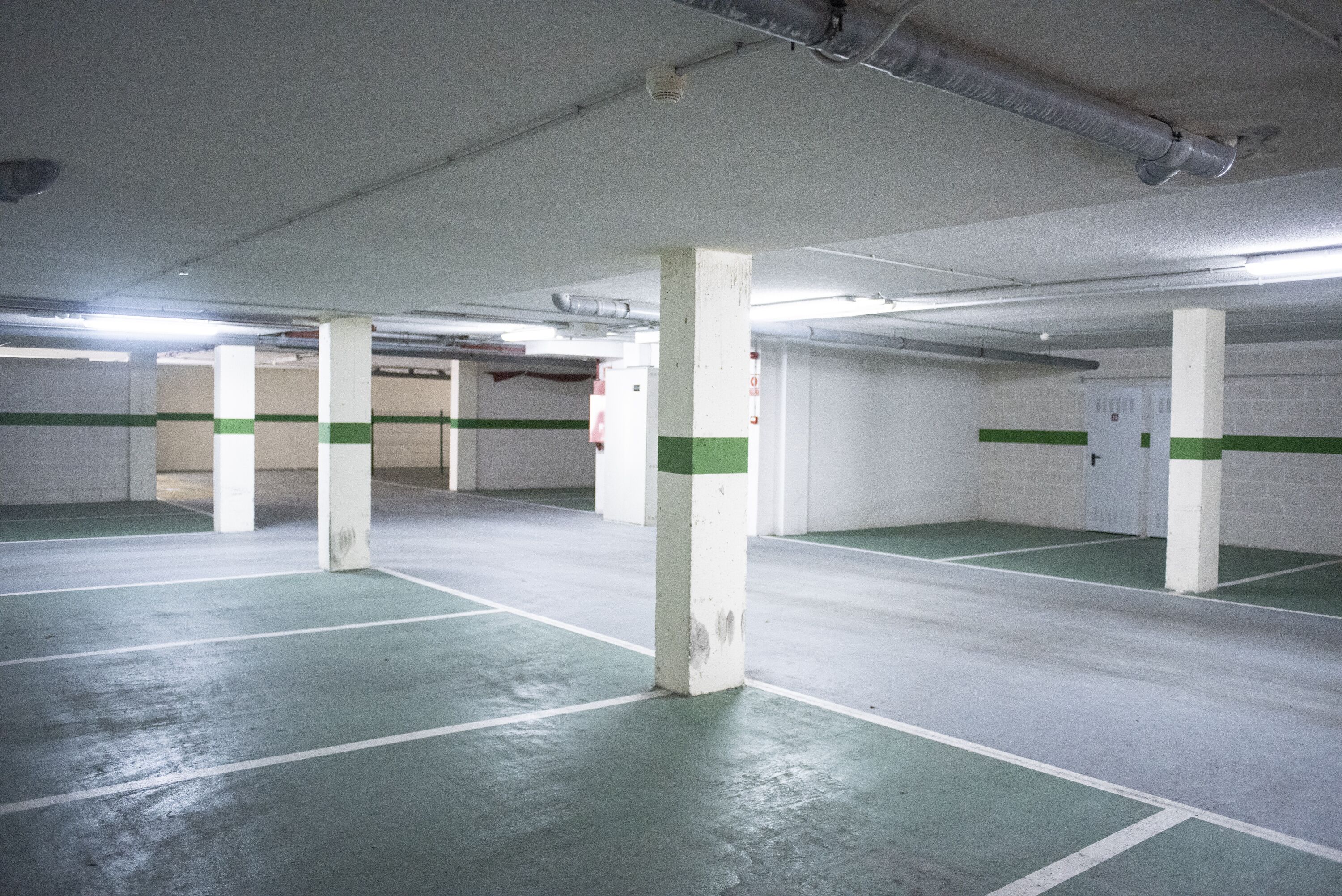An indoor parking garage with white pillars and green stripes, fluorescent lighting, and a concrete floor.
