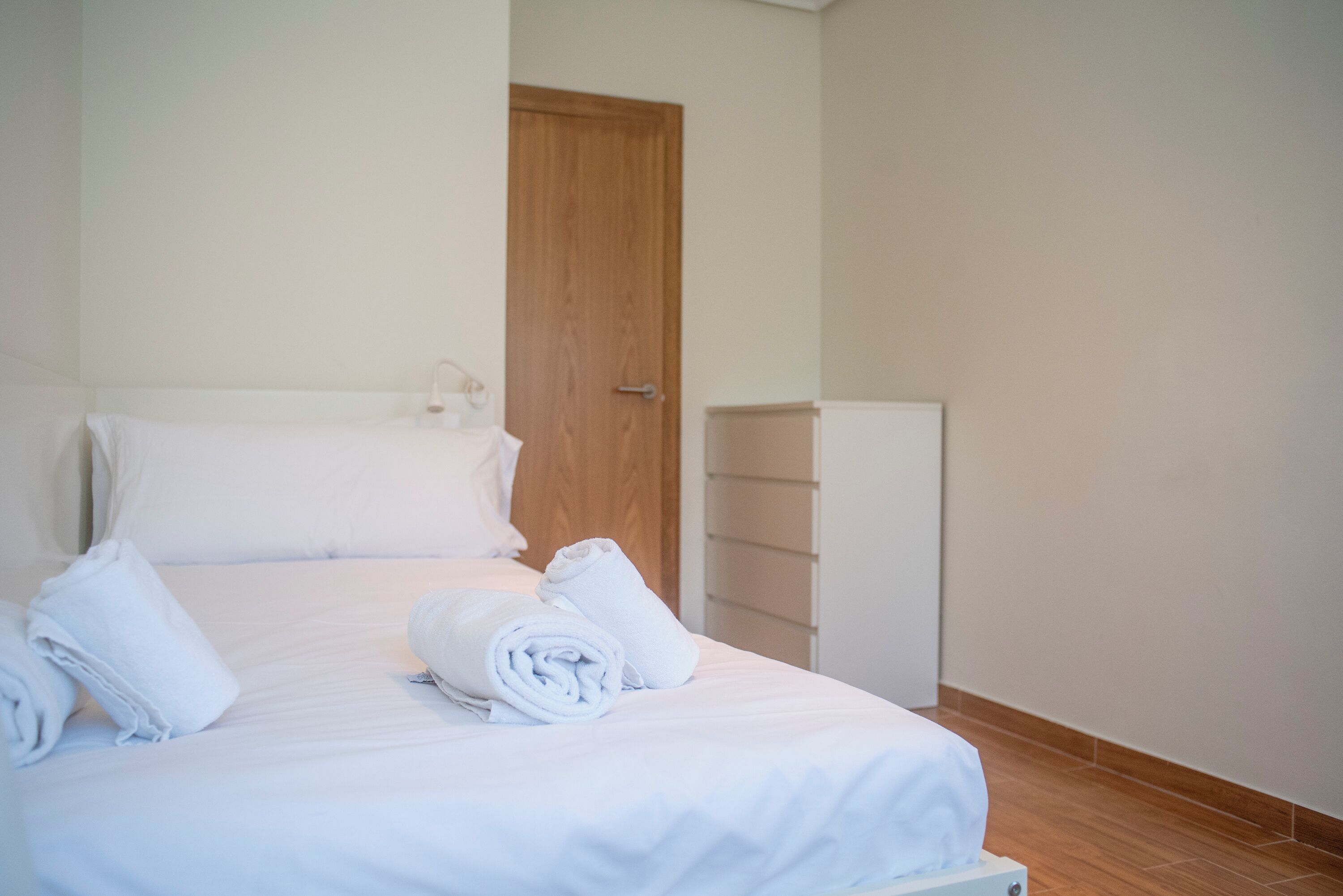 Una cama bien tendida con sábanas blancas y toallas enrolladas, una puerta de madera y un armario blanco.