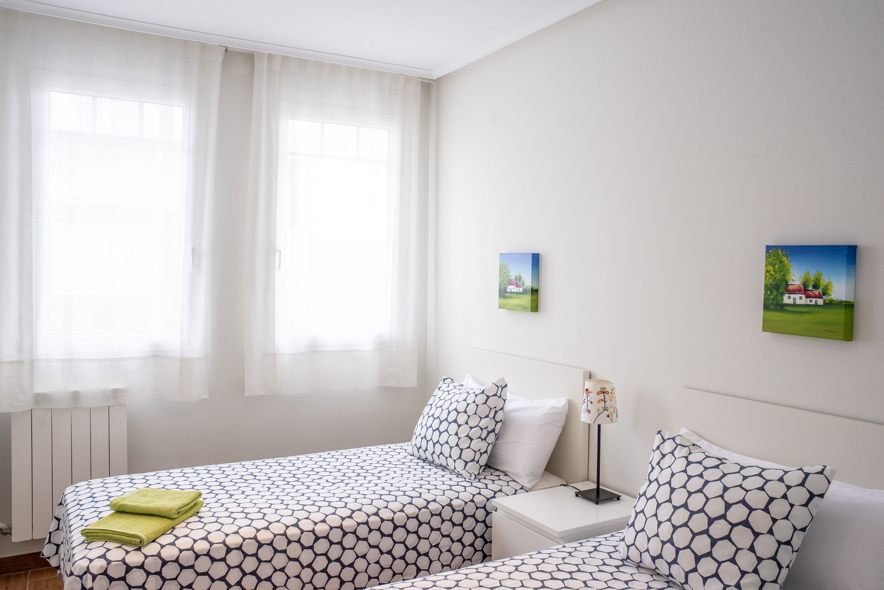 Habitación de hotel con dos camas, una mesita de noche, una lámpara y dos cuadros pequeños en la pared.