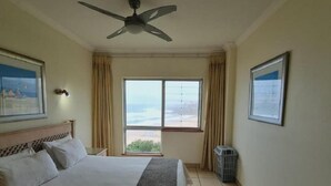 Habitación de hotel con cama, ventilador de techo, ventana con vistas, cubo de basura y un cuadro en la pared.