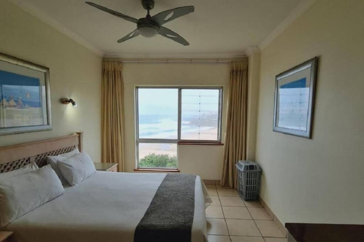 A hotel room with a bed, a ceiling fan, a window with a view, a trash bin, and a framed picture on the wall.