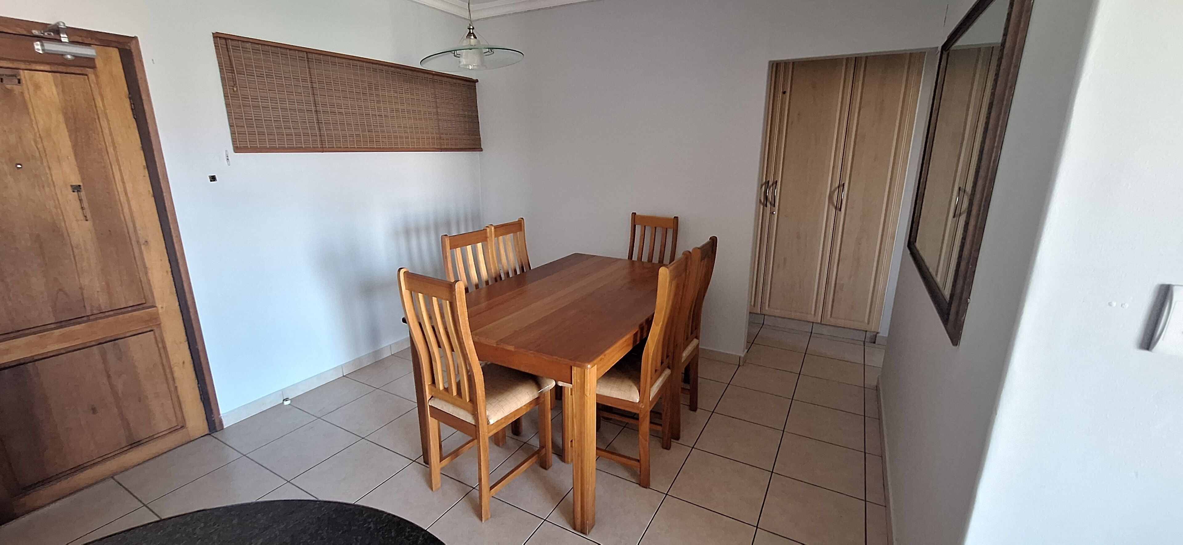 A dining area with a wooden table and chairs, a ceiling light, a window with blinds, and a wardrobe.