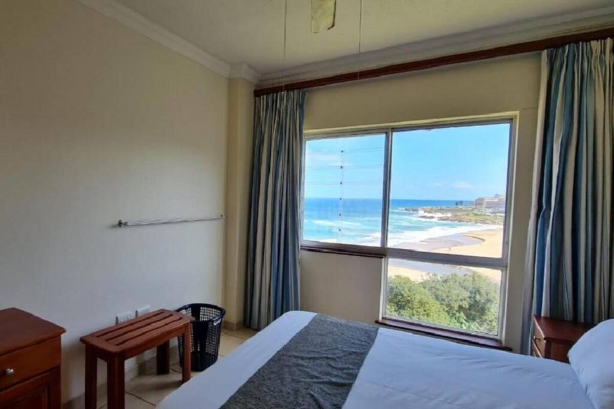 A hotel room with a large bed, a wooden bench, a small table, and a view of the ocean.
