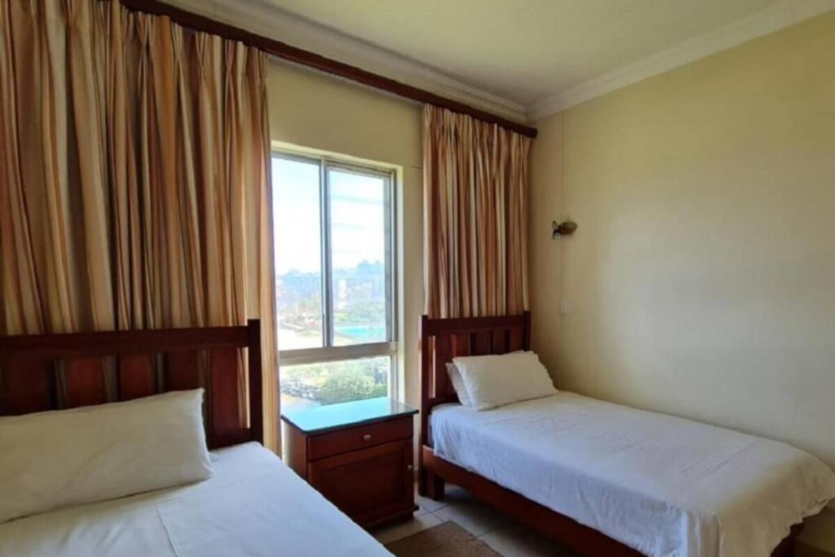 A hotel room with two single beds, a nightstand, a window with curtains, and a view of the outdoors.