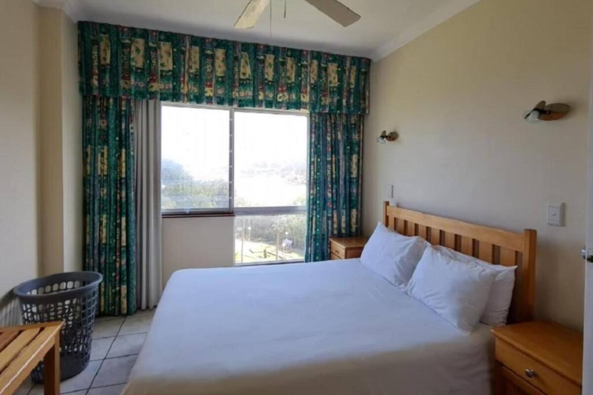 A bedroom with a bed, wooden headboard, a ceiling fan, a window with curtains, a nightstand, and a small table.