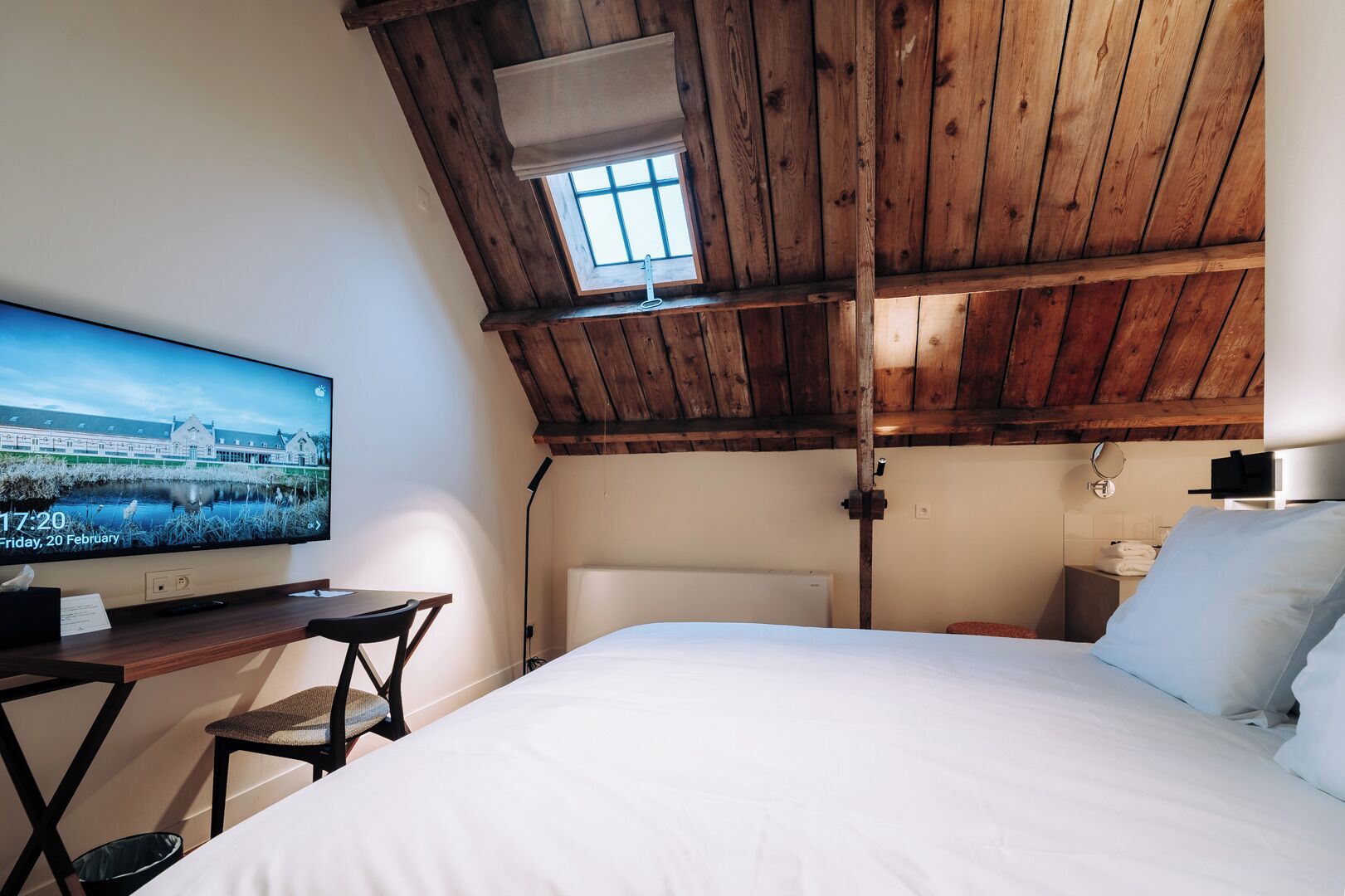 Een kamer met een bed, een bureau met een computer, een raam en een houten plafond.
