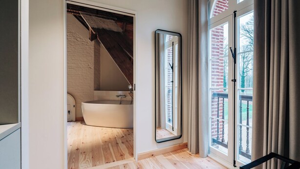 Ein modernes Badezimmer mit einer freistehenden Badewanne, einem großen Spiegel und Blick auf ein Backsteinhaus durch ein Fenster.