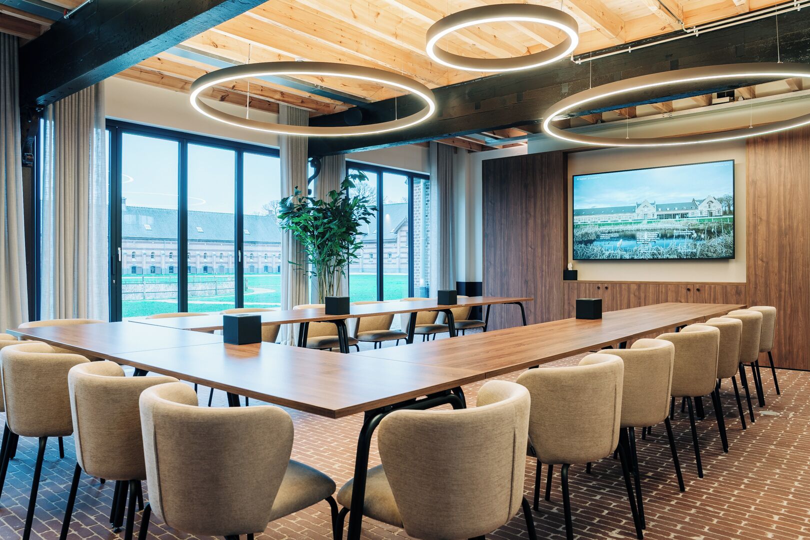 A conference room with a long table, chairs, a large window, and a wall-mounted screen.