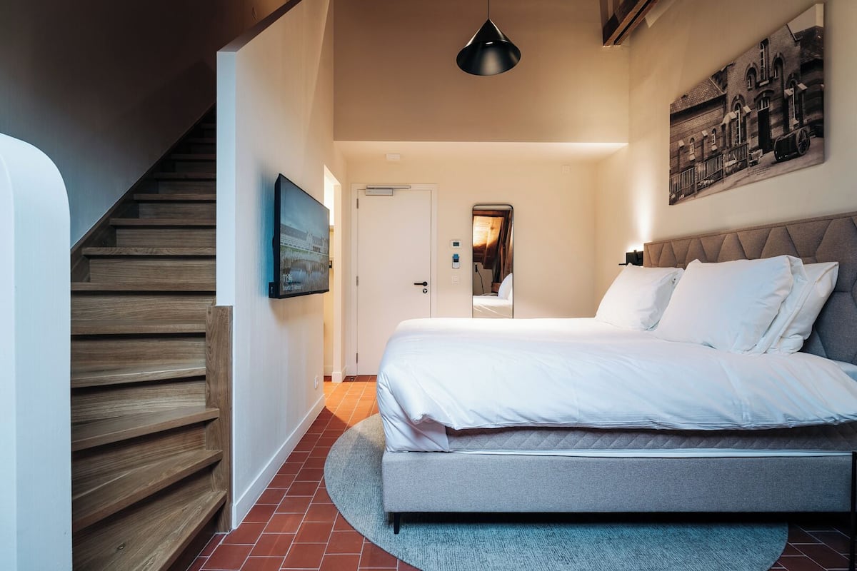 A bedroom with a bed, a wooden staircase, a door, and a wall-mounted picture.