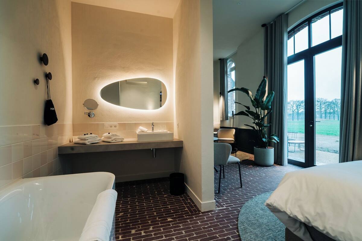 A modern bathroom with a bathtub, a sink, and a large mirror. There is a potted plant and a chair near the window.