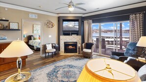 A spacious living room with a fireplace, a ceiling fan, and a view of a body of water through large windows. - Heavenly Hideaway - 2 Pools, Hot Tub, & More! (Lake Ozark)