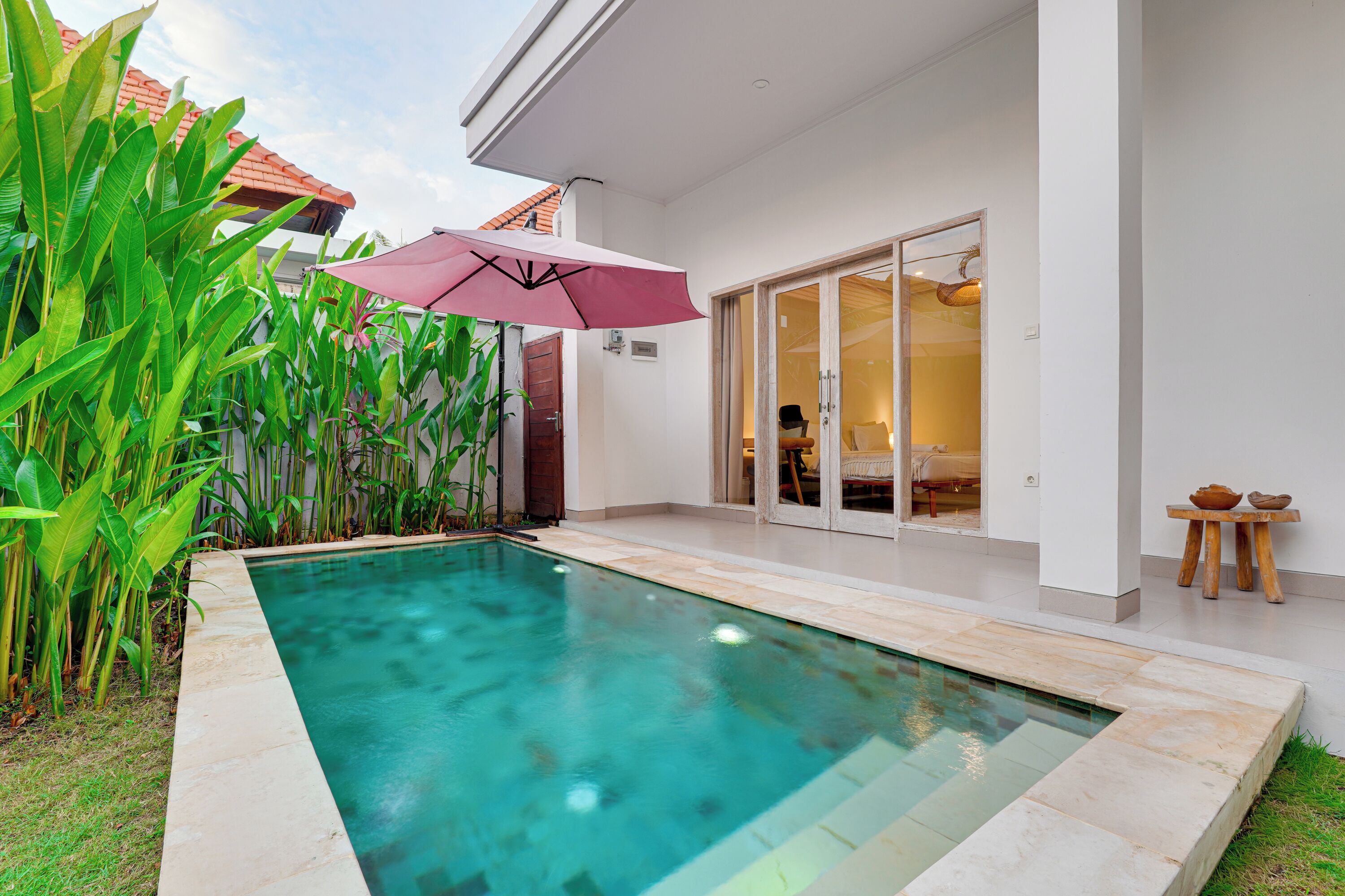 A swimming pool with a tiled deck, a glass door entrance, and a patio area with plants and a table.
