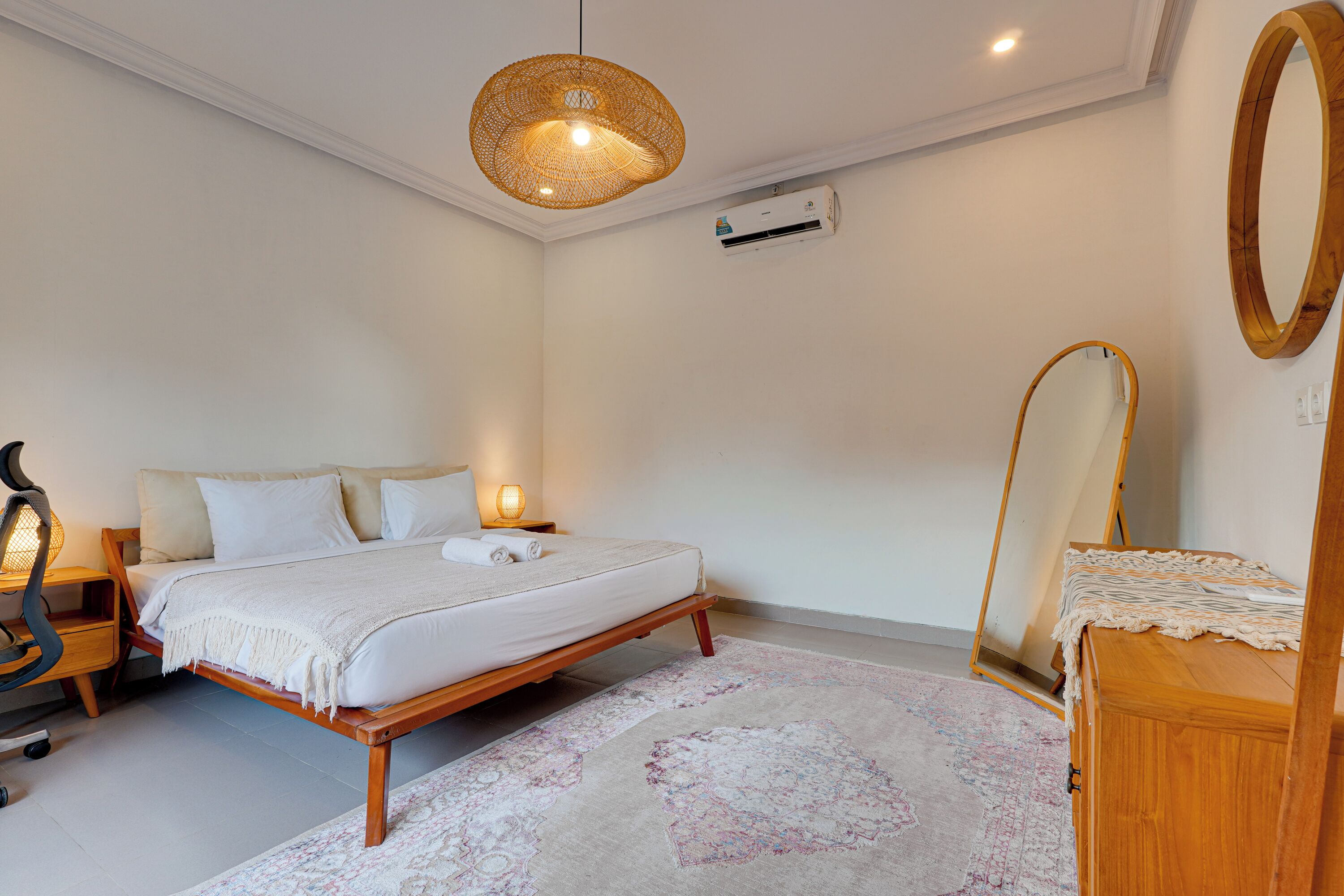 A bedroom with a bed, bedside lamp, wooden nightstand, mirror, and an air conditioning unit.