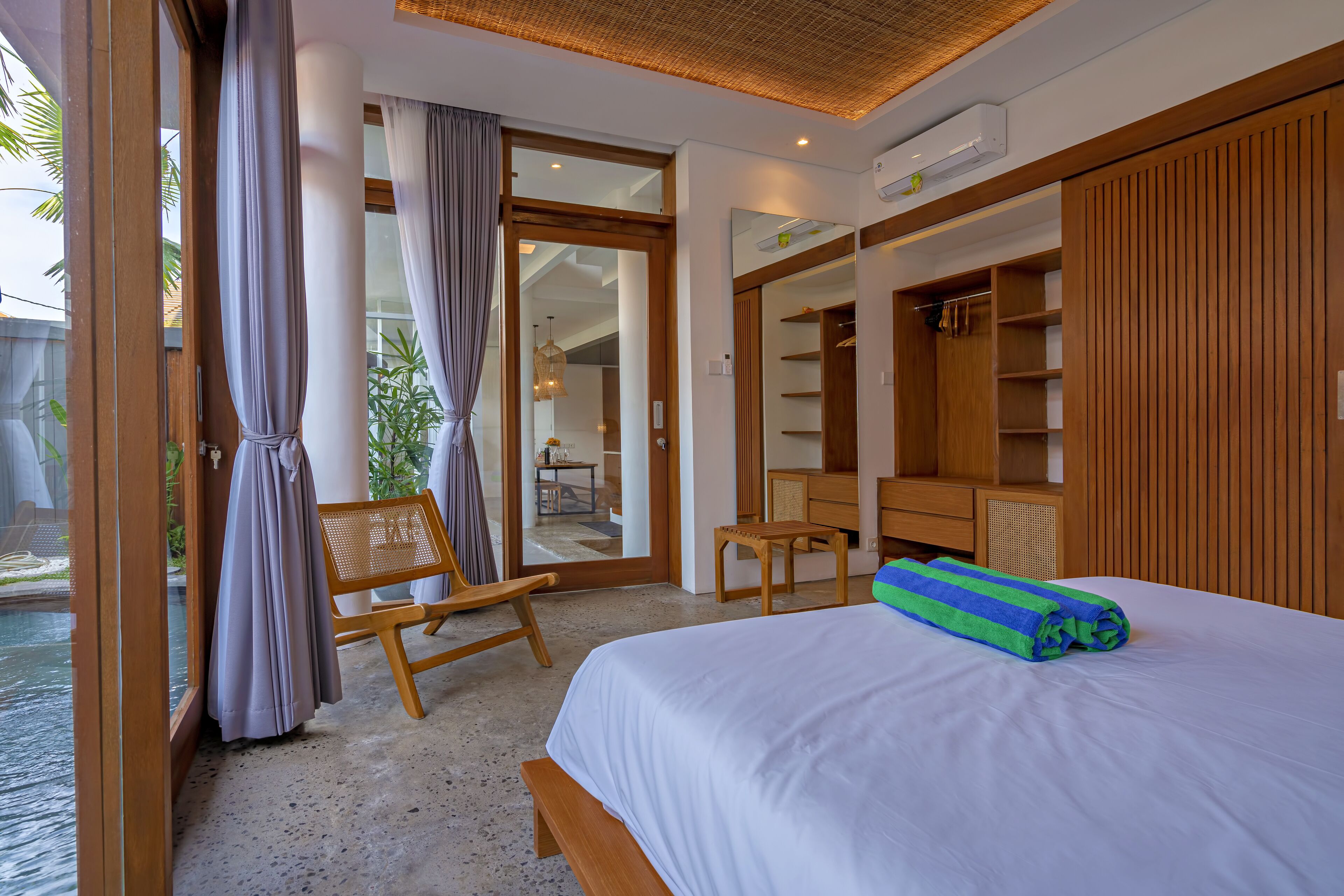 A hotel room with a bed, a bench, a small table, and a view of a pool.