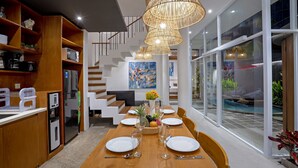 Un espace repas moderne avec une table en bois disposée pour six personnes, entourée de chaises blanches. Une grande fenêtre offre une vue sur une piscine extérieure et sur la verdure.