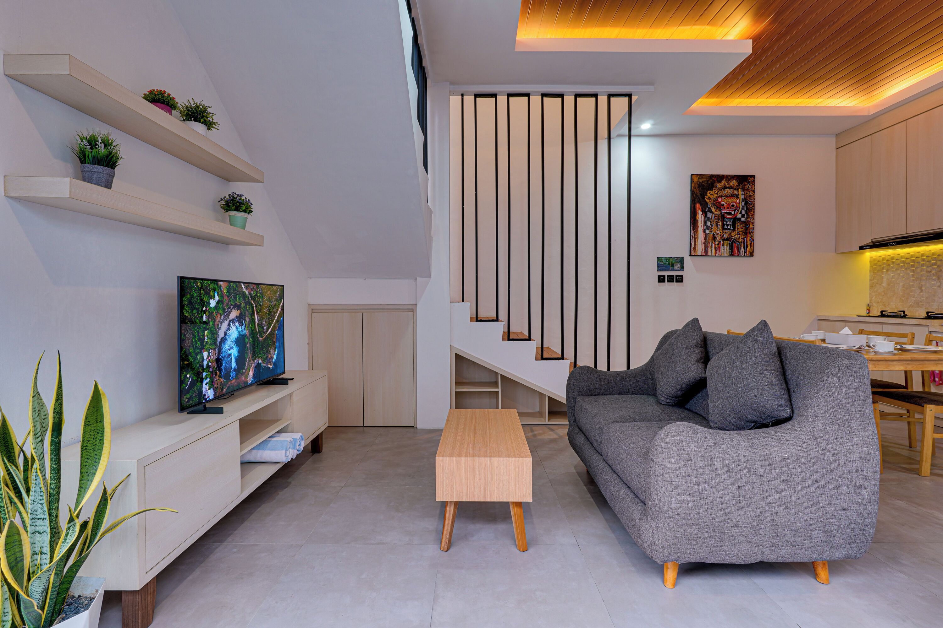 A modern living room with a flat-screen TV, a grey sofa, a wooden coffee table, and a dining area with a wooden table and chairs.