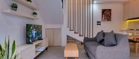 A modern living room with a flat-screen TV, a grey sofa, a wooden coffee table, and a dining area with a wooden table and chairs.