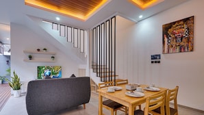 Un comedor moderno con una mesa de madera, sillas y un sofá gris. Una escalera con barandales negros conduce a un nivel superior.