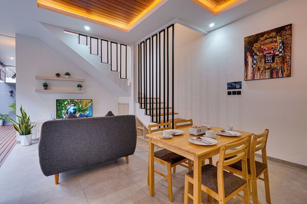 A modern dining area with a wooden table, chairs, and a grey sofa. A staircase with black railings leads to an upper level.