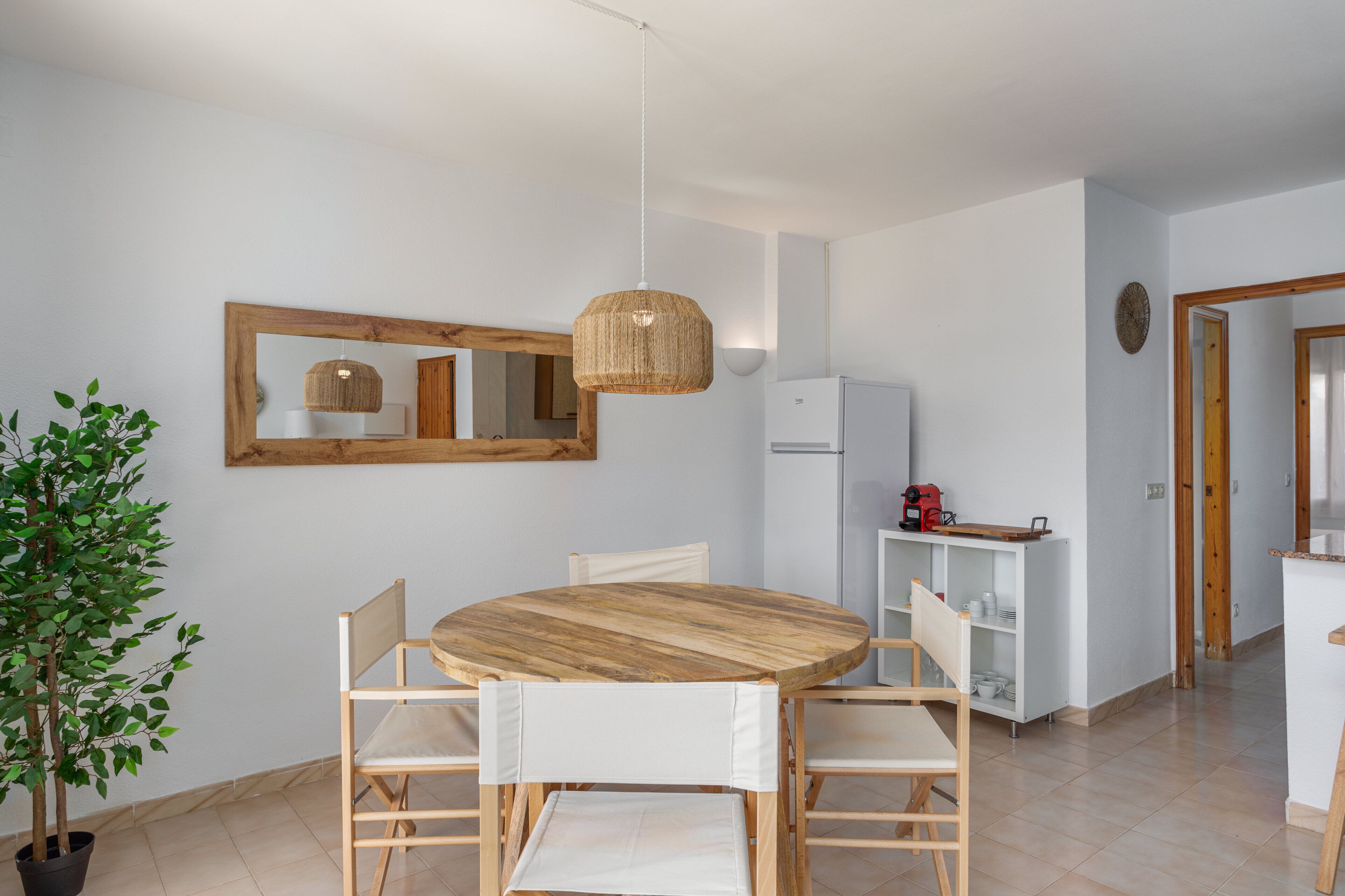A modern kitchen with a round wooden table, white chairs, a hanging light fixture, and a potted plant.
