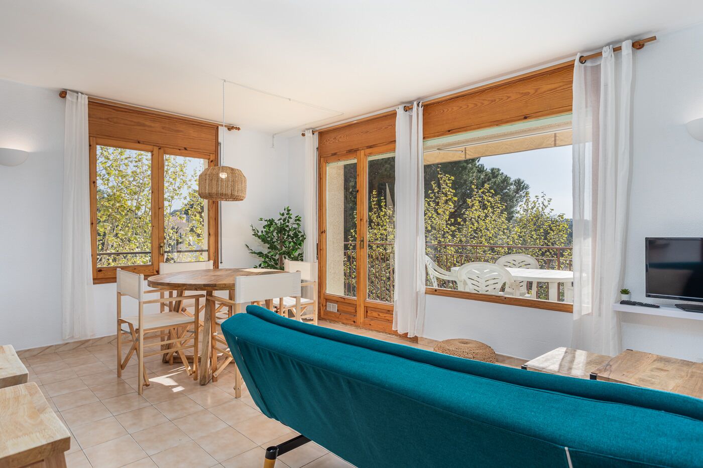 A spacious room with a dining area, a sofa, and a TV. Large windows offer views of greenery and a balcony.