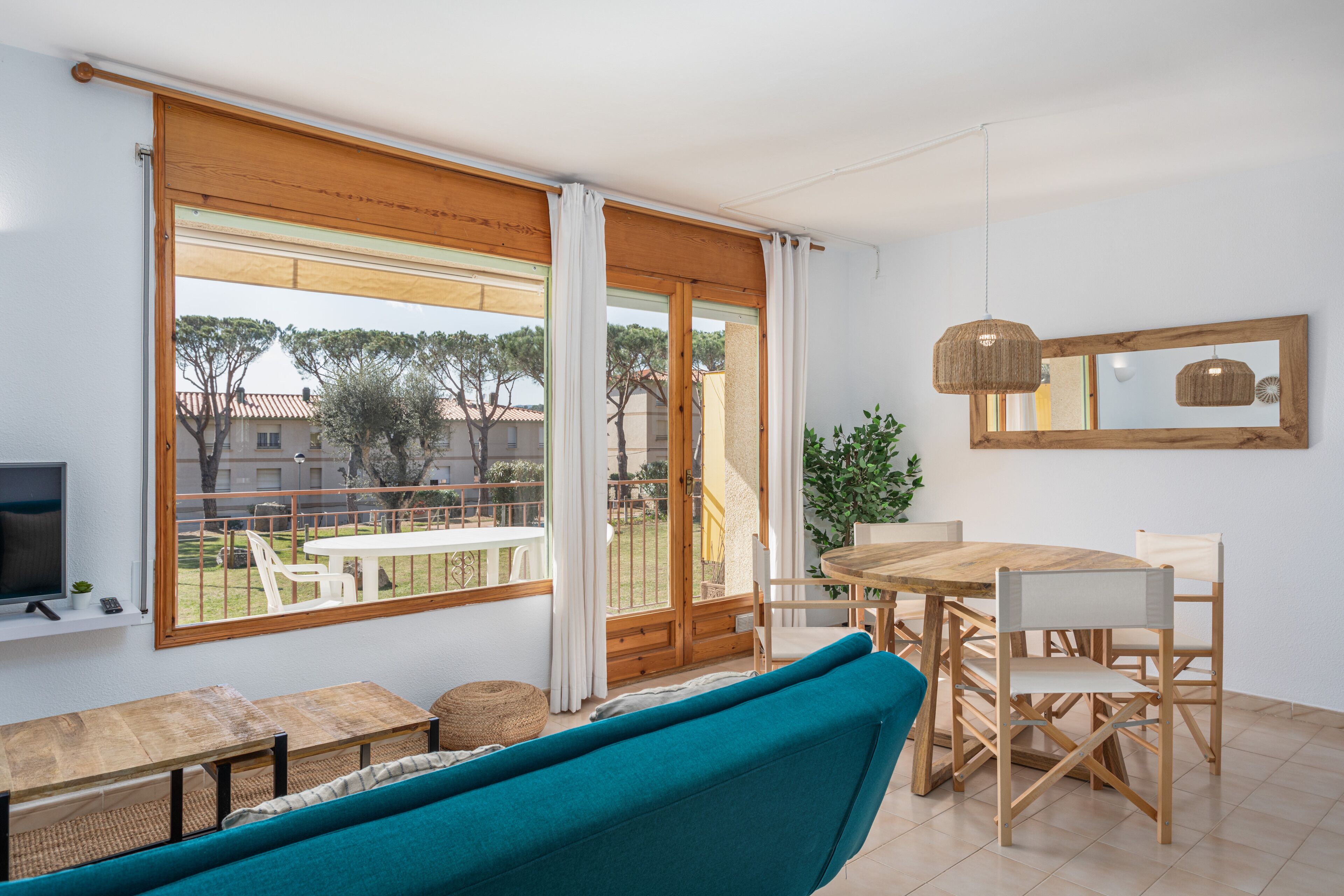 A modern living room with a large sliding glass door leading to a balcony, a wooden dining table with chairs, a blue sofa, and a television on a shelf.