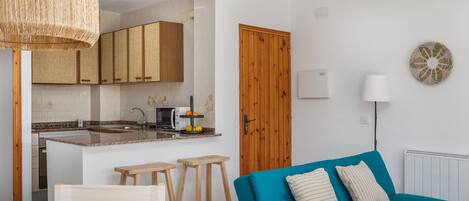A modern kitchen with wooden cabinets, a dining table, and stools. There is a teal sofa with pillows in a living area.