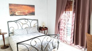 A bedroom with a metal bed frame, a bedside table with a lamp, a view of a sunset through curtains, and a framed picture on the wall. - Room with private bathroom - Brown (Napoli)