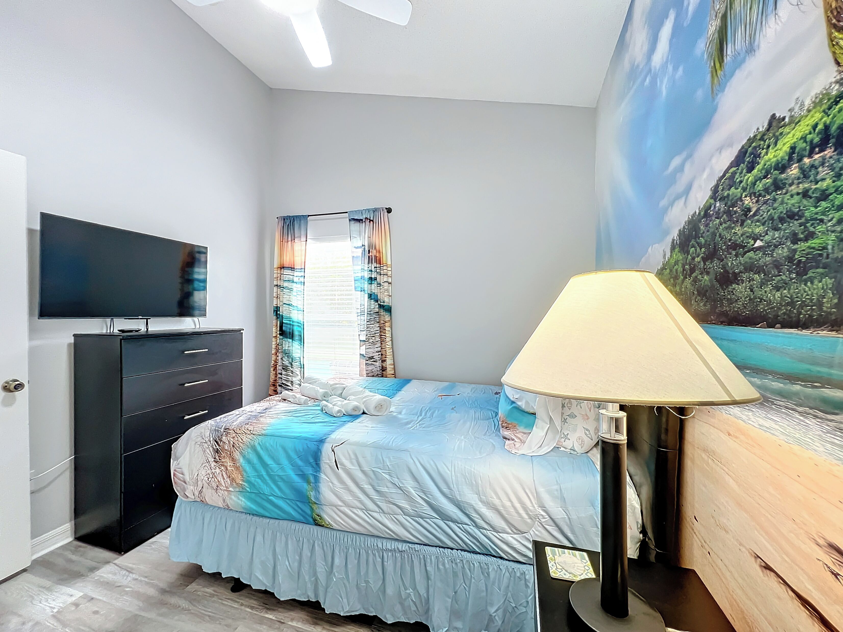 Una camera da letto con un letto, una televisione, una cassettiera e un quadro raffigurante una spiaggia.
