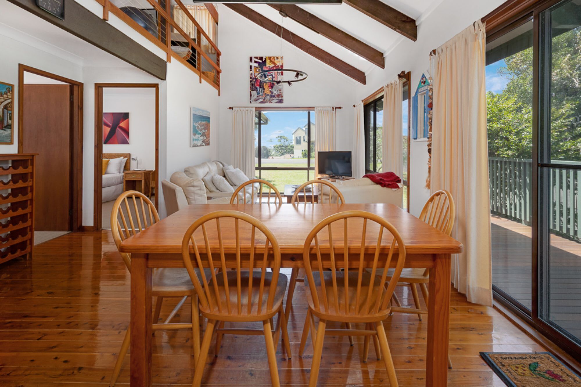 Een ruime woonkamer met een houten eettafel en stoelen, een bank en een televisie. Er is een balkon met uitzicht op de omgeving.