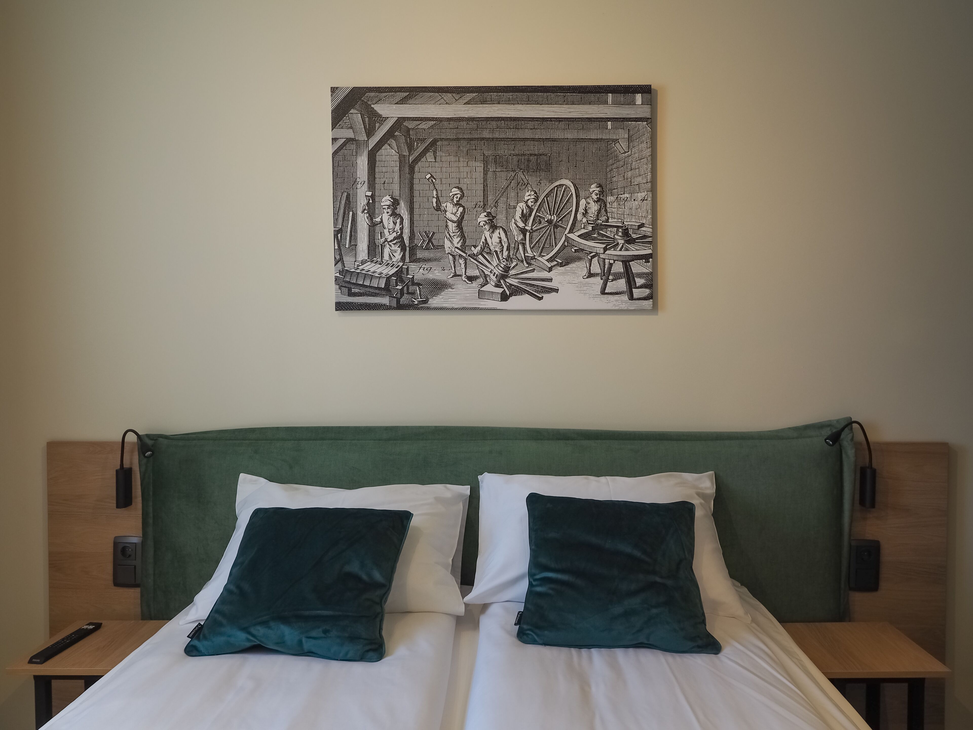 Une chambre d’hôtel avec un lit, deux oreillers, une télécommande et un tableau représentant une scène industrielle, accroché au mur.