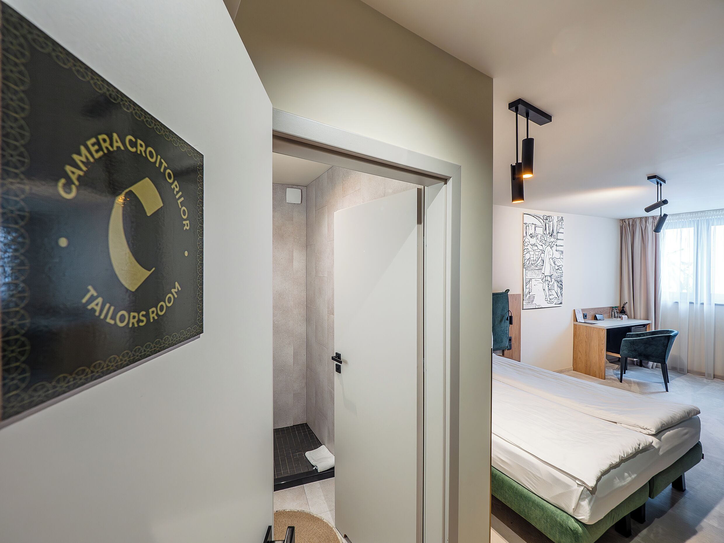 Habitación de hotel con cama, escritorio, silla y ducha, con un cartel que dice 'Tailors Room'.