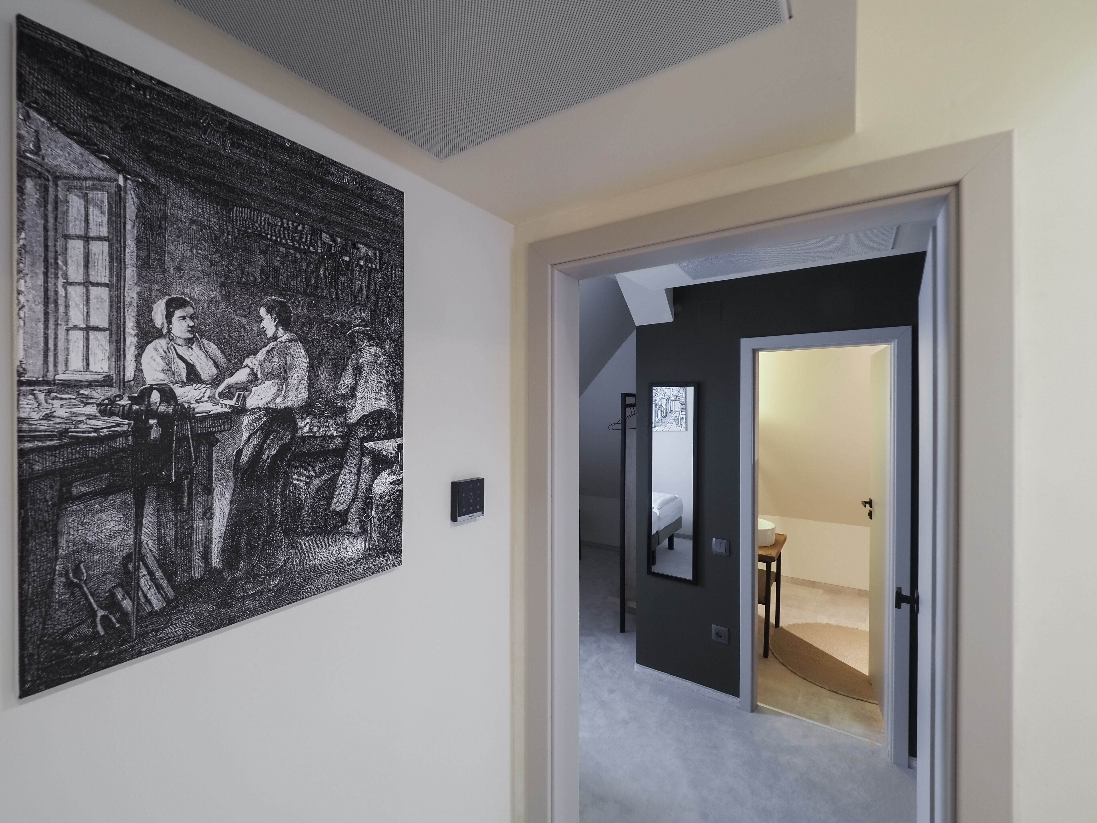 Un pasillo con una obra de arte grande en blanco y negro a la izquierda, una lámpara moderna y una puerta que da a una habitación con cama y una mesita.