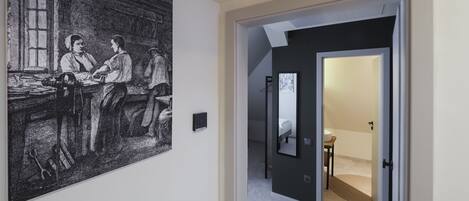 Un pasillo con una obra de arte grande en blanco y negro a la izquierda, una lámpara moderna y una puerta que da a una habitación con cama y una mesita.