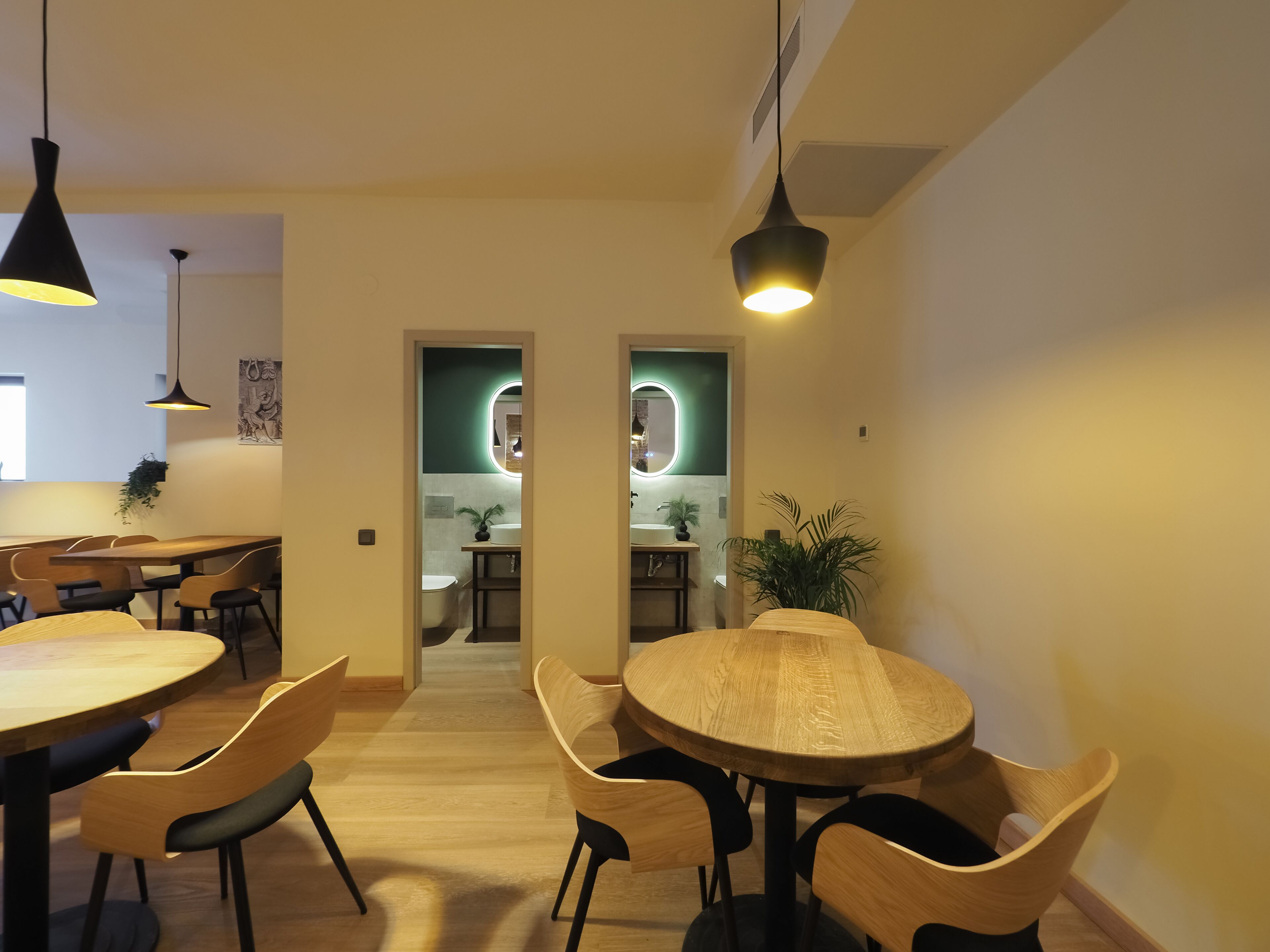 Un comedor moderno con mesas y sillas de madera, iluminación colgante y vistas hacia un baño con una pared verde de acento.