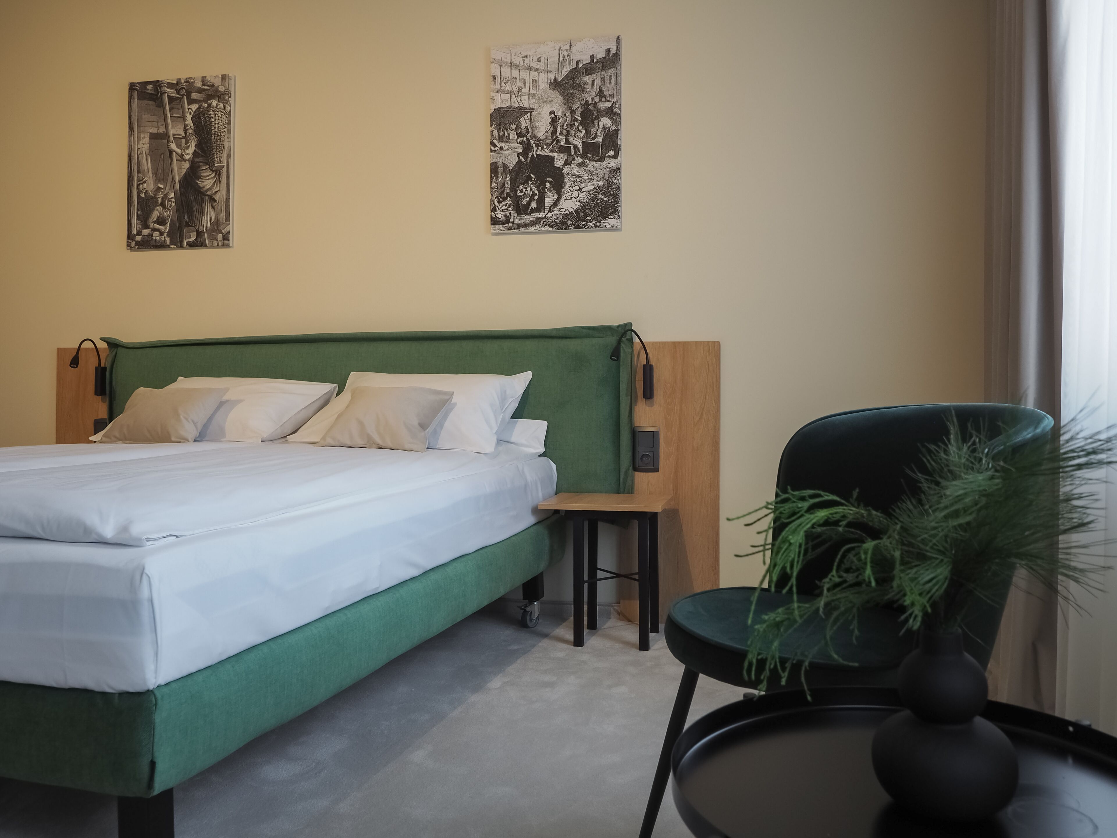 Cama bem arrumada com roupa de cama branca, cabeceira estofada verde e uma pequena mesa de cabeceira com abajur. Há uma cadeira com uma planta ao lado da cama. Duas fotografias em preto e branco estão penduradas na parede acima da cama.