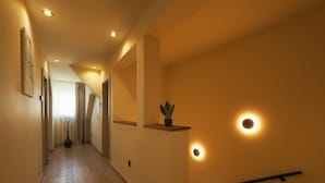 Um corredor com piso de madeira, luzes embutidas no teto, uma planta em vaso e um corrimão no lado direito.