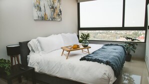 Una camera da letto moderna con un letto grande, un comodino con una lampada, un vassoio con prodotti per la colazione e vista sulla città dalla finestra.