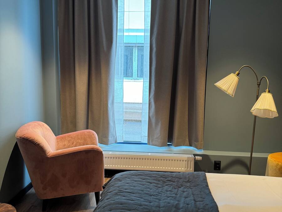Una habitació d'hotel amb un llit, una cadira, un escriptori, una làmpada i una finestra amb cortines.